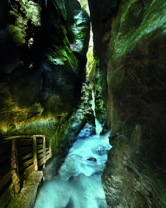 Schmale Schlucht mit Holzsteg und türkisfarbenem Fluss zwischen steilen moosbedeckten Felsen